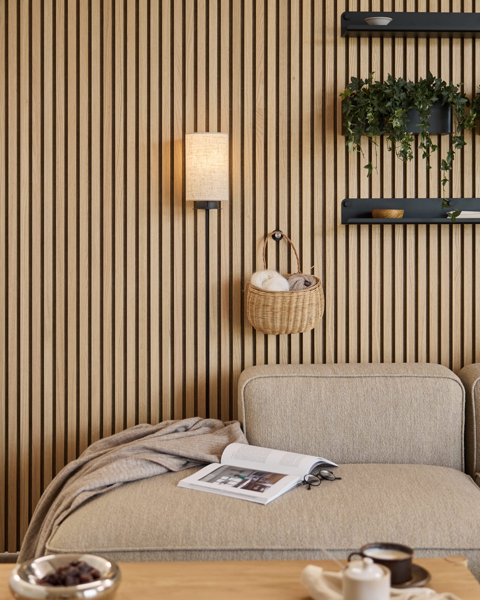 Beige sofa with blanket and book beside a WoodUpp Akupanel acoustic slat wall, shelves with plants and decor, and a Feya Wall Lamp in Black/Beige.